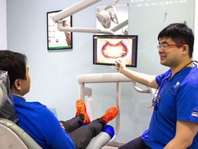 A dentist shows a patient their digital mouth scan on a monitor while discussing treatment steps related to a full denture implant.